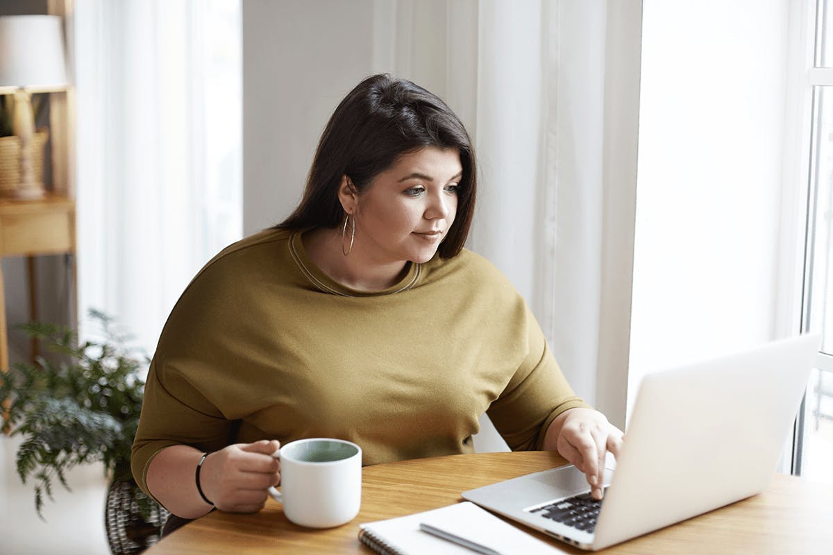 Kvinna sitter framför datorn, dricker kaffe och läser