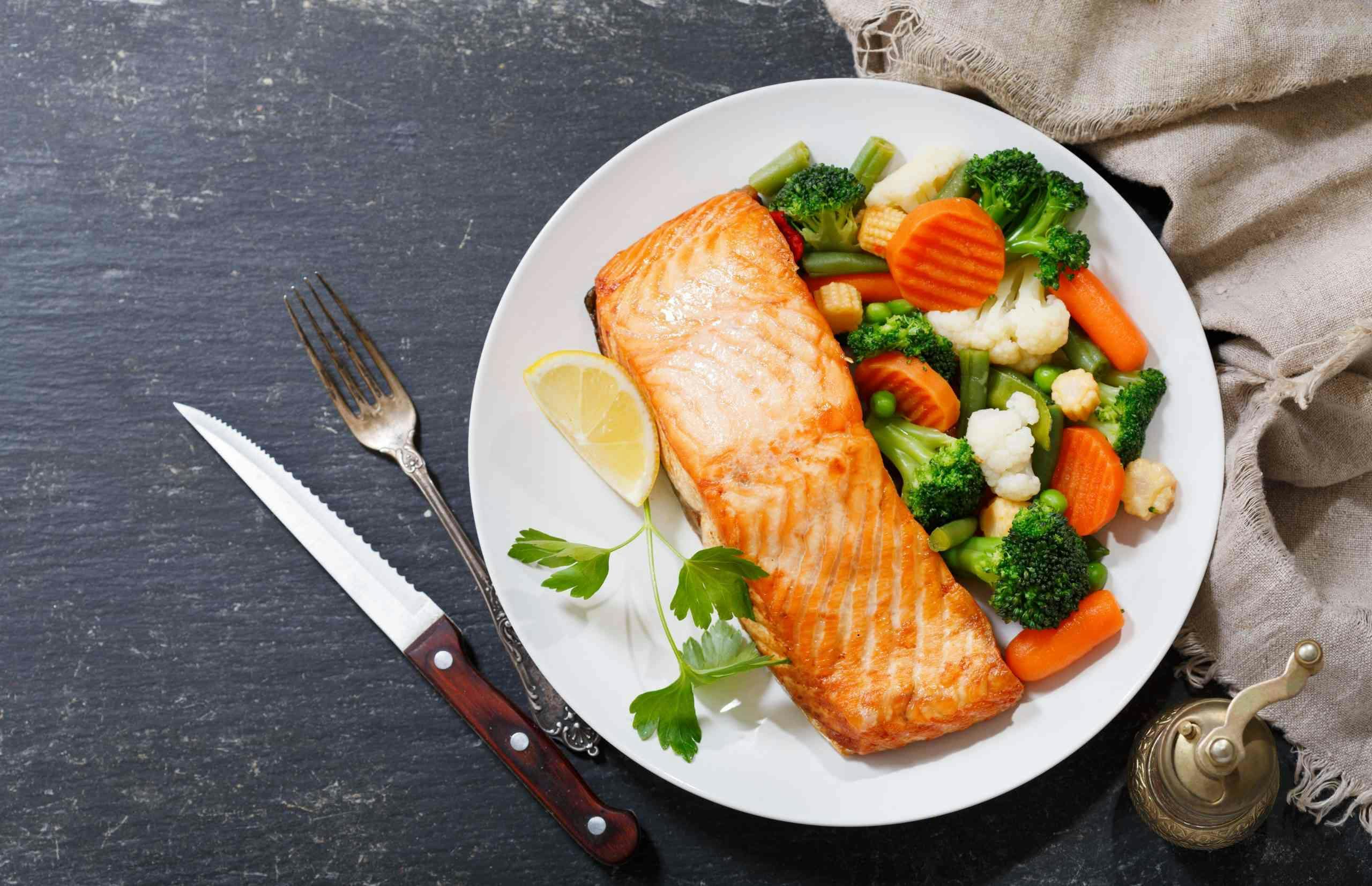 <p>plate of baked salmon steak with vegetables on dark background, top view</p>
